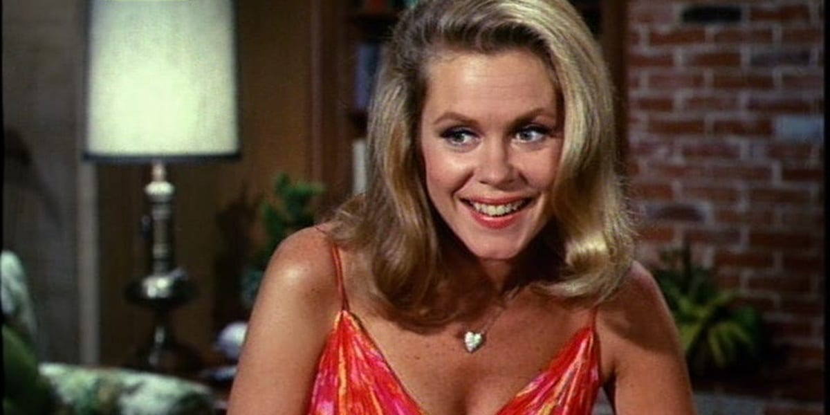 Samantha smiling and looking to her right on Bewitched, wearing a heart necklace and an orange-red tank top or dress with a living room in the background