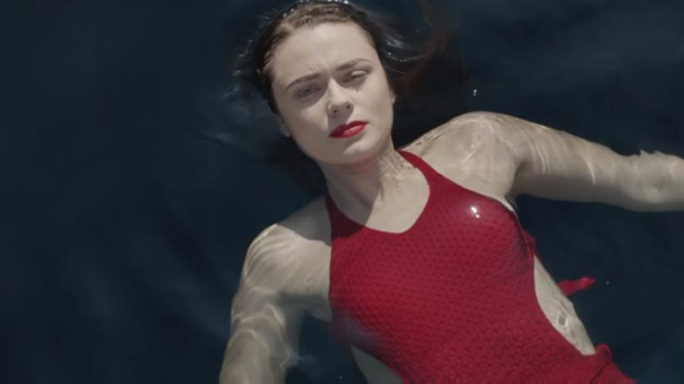 Vera is lying on her back in the sea. She looks reflective and is wearing a red swimming costume with cut out sides