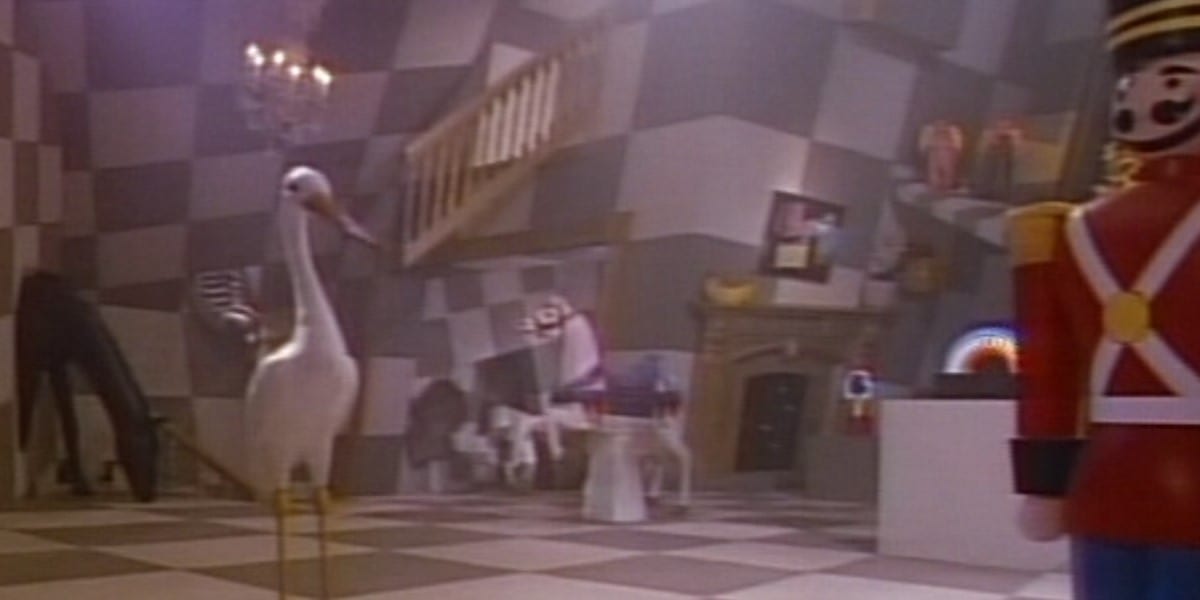 toys of a stork and a life-size nutcracker stand in the foreground of a black and white checkerboard room with warped corners and a staircase to nowhere over a warped fireplace. More toys also appear in the background.