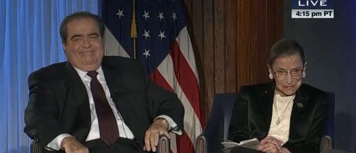 Justice Antonin Scalia and Justice Ruth Bader Ginsburg sitting in chairs with an American flag behind them.