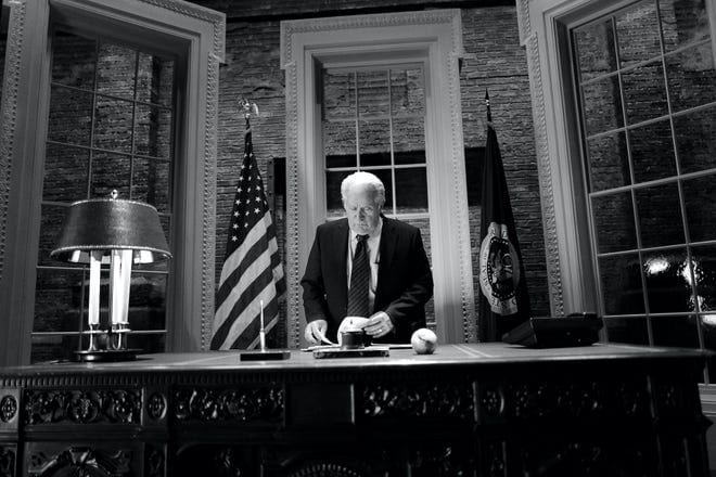 Martin Sheen as an older Jed Bartlet stands behind the desk in the Oval Office in a black and white shot