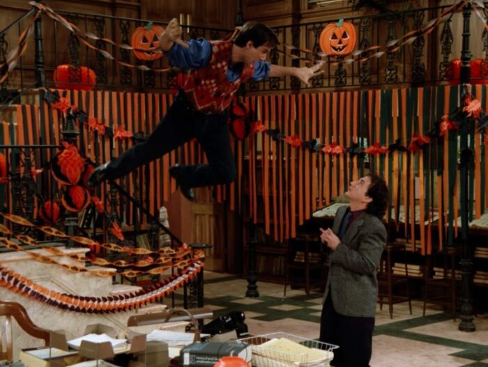 Balki floats above Larry, his arms and legs flailing around a heavily halloween decorated office, Larry looks frightened underneath