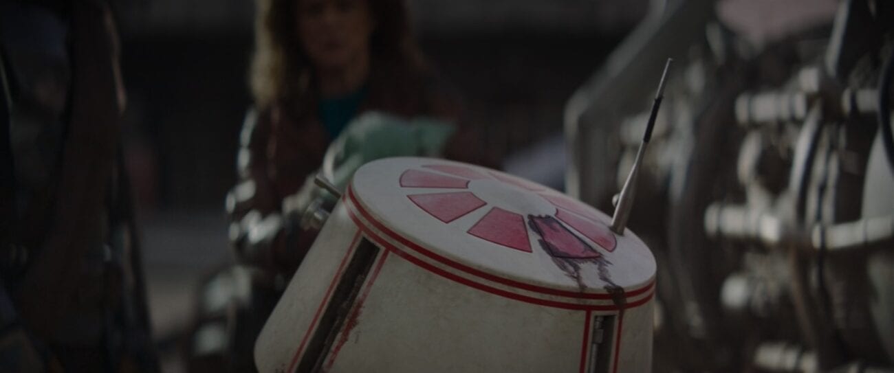 R5-D4 rolls towards The Mandalorian, Peli Motto and The Child, showing damage on his dome