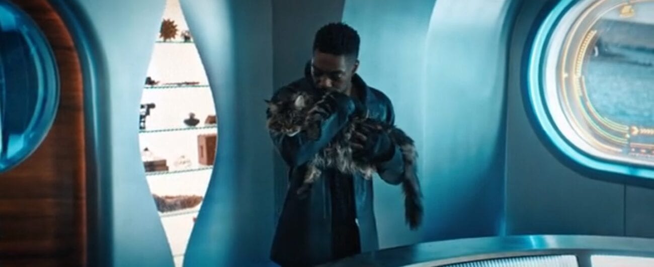 Book (David Ajala) stand in the interior of his spaceship holding his large stripped cat, Grudge, in both arms close to his face, with a shelf of space knicknacks and an oval window in the backgroun
