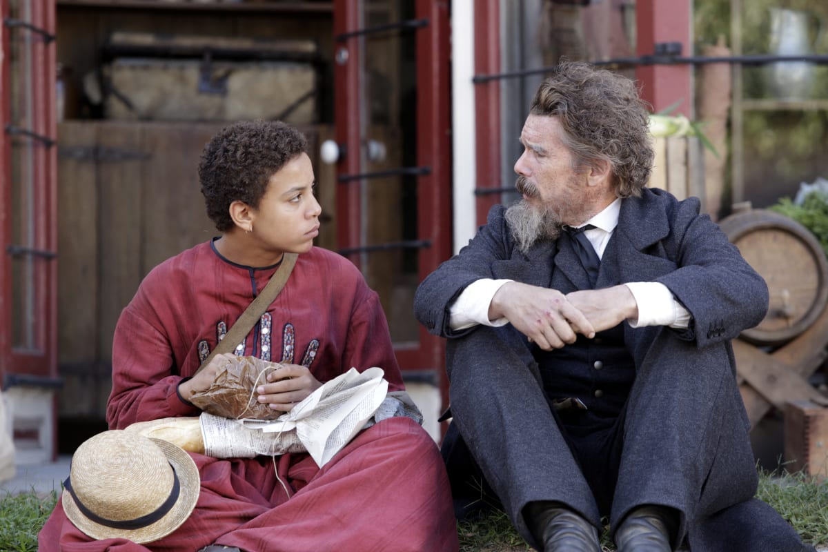 Onion (Joshua Caleb Johnson) in a red dress with a hat and parcel in his lap and John Brown (Ethan Hawke) with his arms crossed in front of his knees, sitting on a stoop looking at each other