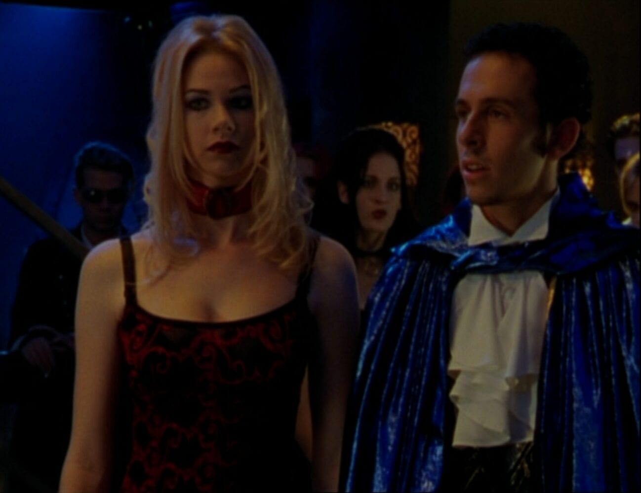 Wearing a dark red dress and a choker, Chanterelle stands in the club with Diego, who wears a ruffled shirt and blue cape, and the rest of the vampire cult, also dressed in vampire attire