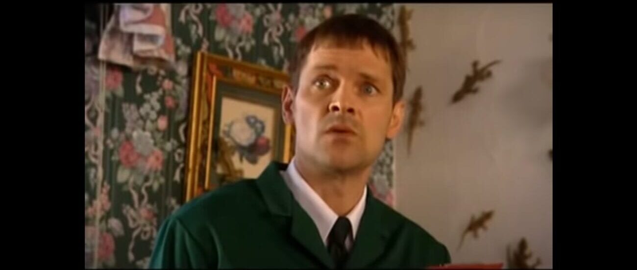 Mark Heap looks on quizzically as he stands in front of a wall with patterned wallpaper and a framed picture