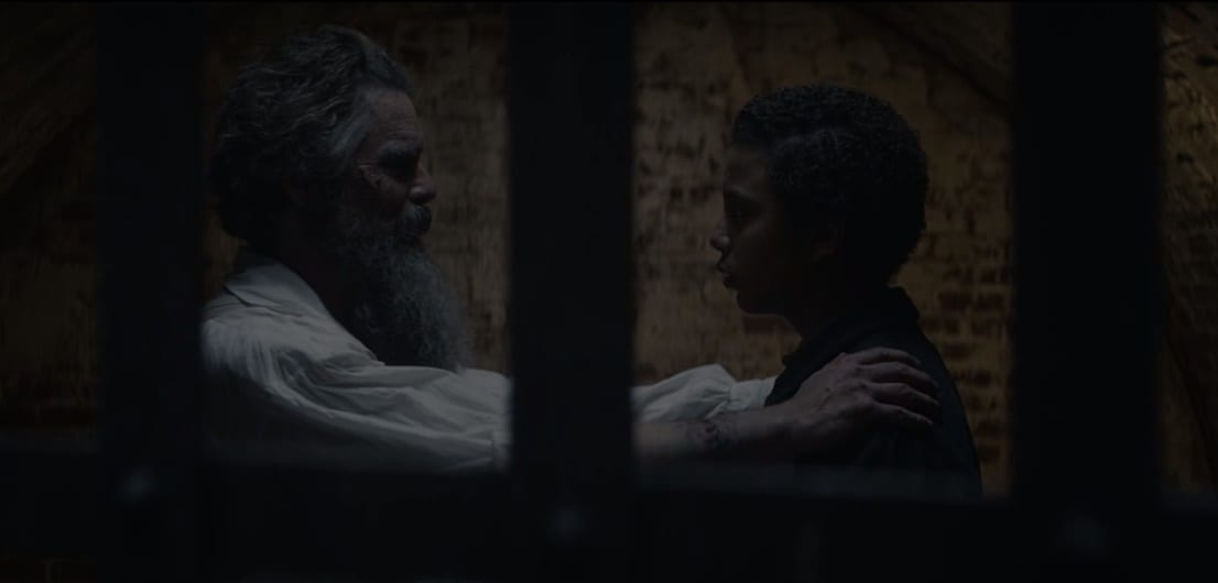 John Brown (Ethan Hawke) with his arms on Onion's (Joshua Caleb Johnson) shoulders in a darkened jail cell seen through the bars