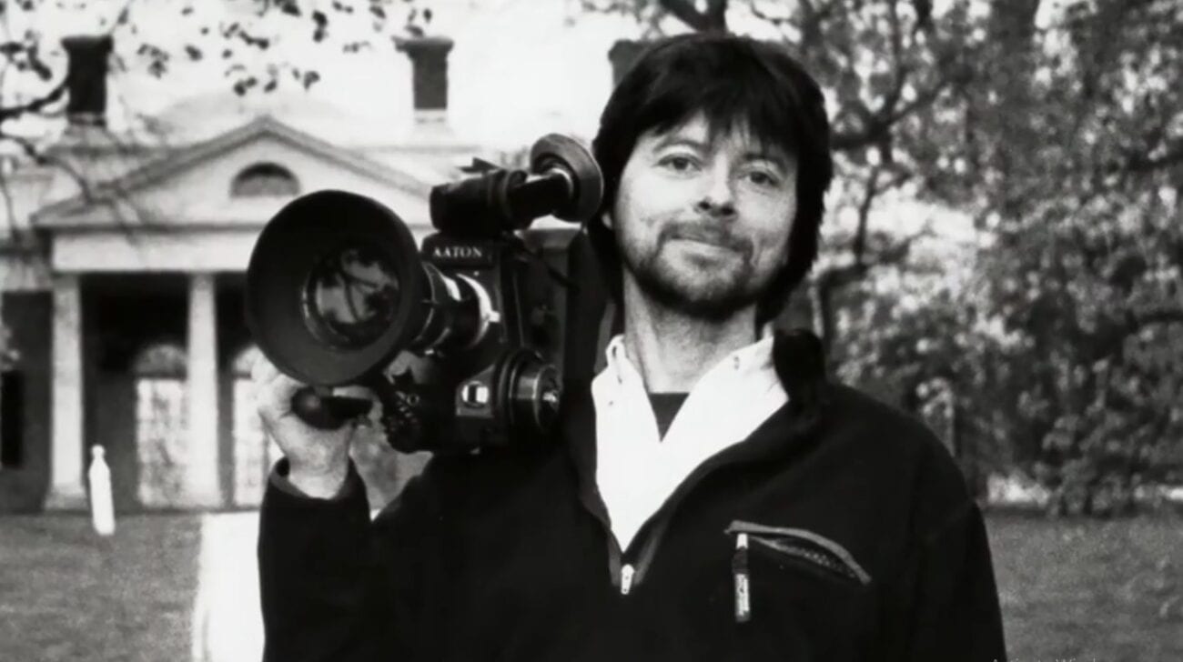 Black and White photo of a young Ken Burns holding a large film camera over his shoulder, standing in front of a white house