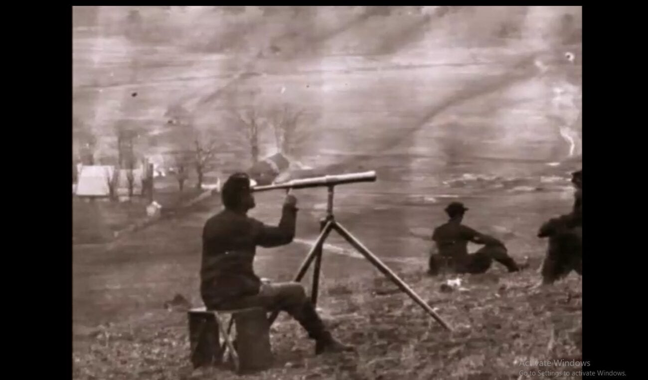 A sepia-toned photograph of a Black soldier sitting at a telescope on a battlefield. Another man sits in front of him on the ground.