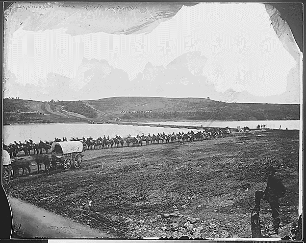 A black and white photograph of an open fields with a column of troops lined up near a river accross the center, a solitary figure in a uniform stands to the right looking at the troops