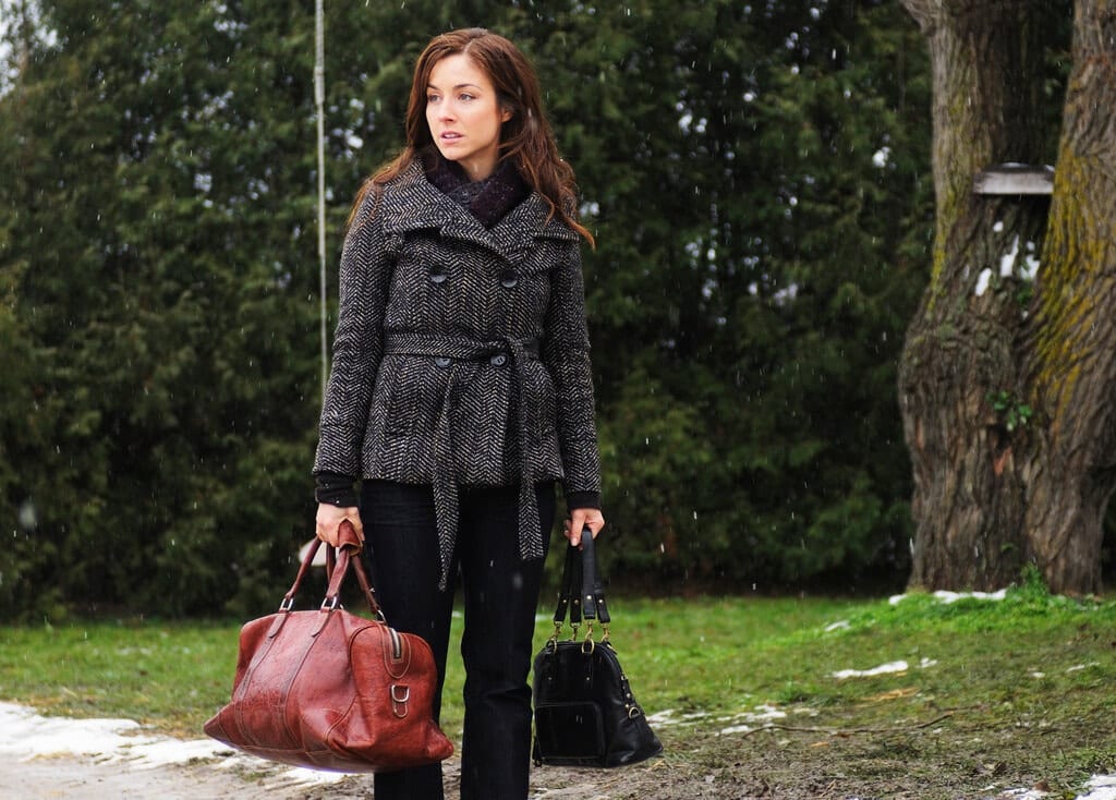 Erica looks offscreen while holding two bags and wearing business casual clothes.