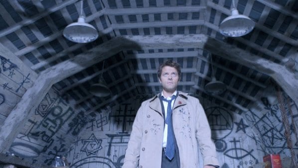 Castiel stands in a trenchcoat and tie in front of a wall covered in symbols