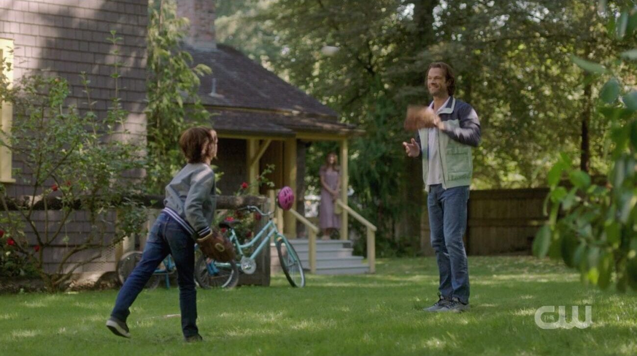 Sam plays catch with his son Dean while blurry blonde woman looks on from the porch in the Supernatural finale