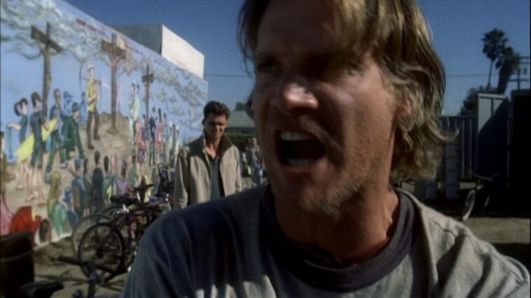 Butchie rants angrily toward the camera as John stands behind him with a mural of three crosses on a hilltop and surfers running toward them on the wall in the background