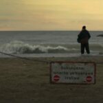 A fisherman stands looking out at the ocean with a sign in Xhosa that reads "sukungena ibhorho yenqanawa ivaliwe" in the foreground