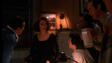 Lana Milford sits in the middle of a darkened office, with the only light behind her on a file cabinet, being surrounded and stared at by the men of the office