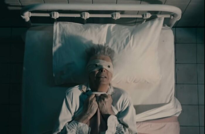 David Bowie in a bed clutching the covers and wearing a button-eyed blindfold from the video for "Lazarus."