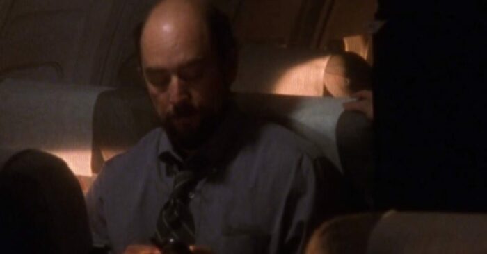 Toby (Richard Schiff) sitting in a plane seat looking at his phone