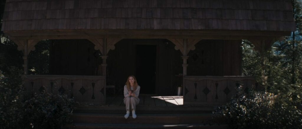 A perfectly straight wood cabin sits on cobblestones in front of a mountain, and Wanda sits on its porch. This is a closer shot, to see Wanda's blank expression better.