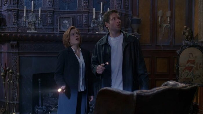 Scully and Mulder are spooked as they stand in front of the mantel in a haunted house