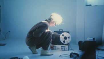 Maryanne Amacher crouches on the ground in front of a tape player and oscillator.