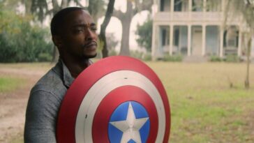 Sam stands holding the Captain America shield in front of him on a lawn