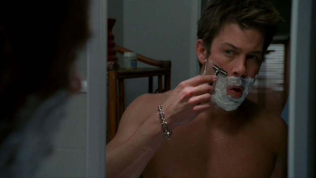 Lindsey, shirtless, looks in the mirror while shaving his face