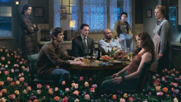 The cast of Six Feet Under sits and stands around a table with flowers in the foreground and a window behind them