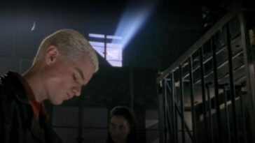 Spike looks downwards as Drusilla stands in the background and sun filters through a warehouse window