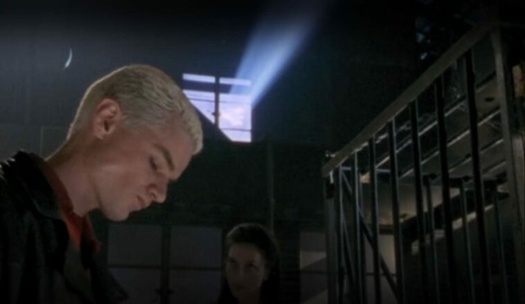 Spike looks downwards as Drusilla stands in the background and sun filters through a warehouse window