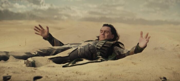 Loki pulls himself out of the sand after crash-landing in the desert.
