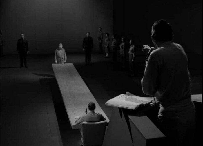 Still from the Twilight Zone episode "The Obsolete Man." Romney Wordsworth stands at the end of a long table while the Chancellor looms over him.