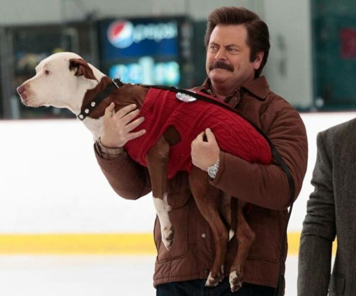 Ron Swanson carrying Champion, who is wearing a sweater, across the ice in a hockey arena