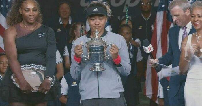 2018 US Open Champion Naomi Osaka holds the trophy as runner-up Serena Williams watches.