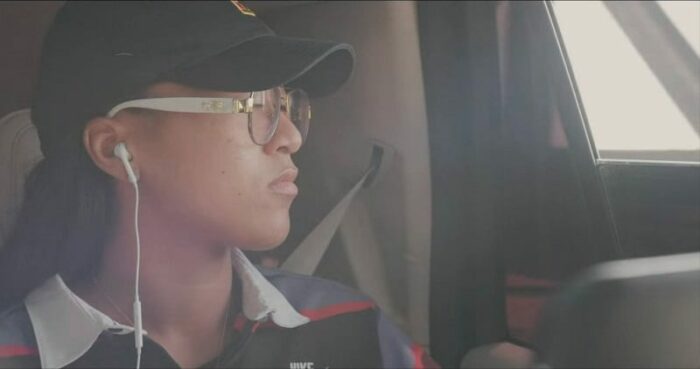 Naomi Osaka is depicted wearing a cap, glasses, and headphones in the back seat of a car.