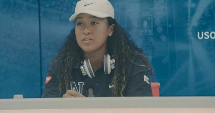 Naomi Osaka is depicted wearing a cap during a post-match press conference.