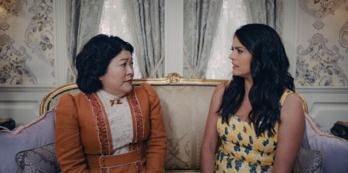 Mrs. Menlove (Ann Harada) and Mel (Cecily Strong) sitting on a couch in the Menlove house looking at each other