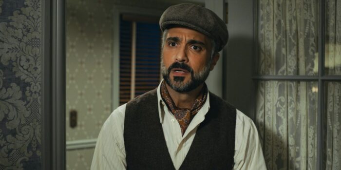 Doc Lopez (Jaime Camil) standing in front of rustic wall, wearing a newsboy cap and looking sad