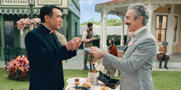 Mayor Menlove (Alan Cumming) stands at a bakesale table with his arms stretched out toward Rev Layton (Fred Armisen) as they share a moment