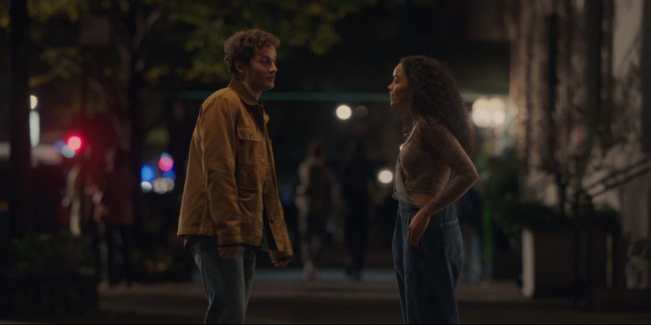 Obie, left, a white boy with curly brown hair wearing a yellowish-brown coat and jeans talking to Zoya on the sidewalk outside of her apartment building at night.