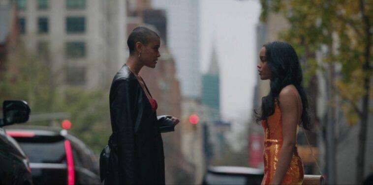 Julien (left) and Zoya (right) standing on a sidewalk on an overcast day. Julien is wearing a red dress covered by a heavy leather coat and has one some gold earrings. Her left arm is facing upward and her fingers are clutched. Zoya is wearing a vibrant sequined orange dress and carrying a sparkling gold purse hung around her shoulder by a gold chain. Her hair is styled with gentle waves. The two are talking and are displeased with one another.