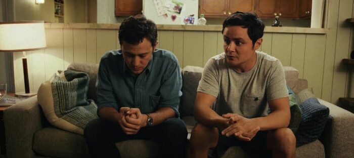 Josh and Victor sit on their couch, each holding his hands clasped in front of him