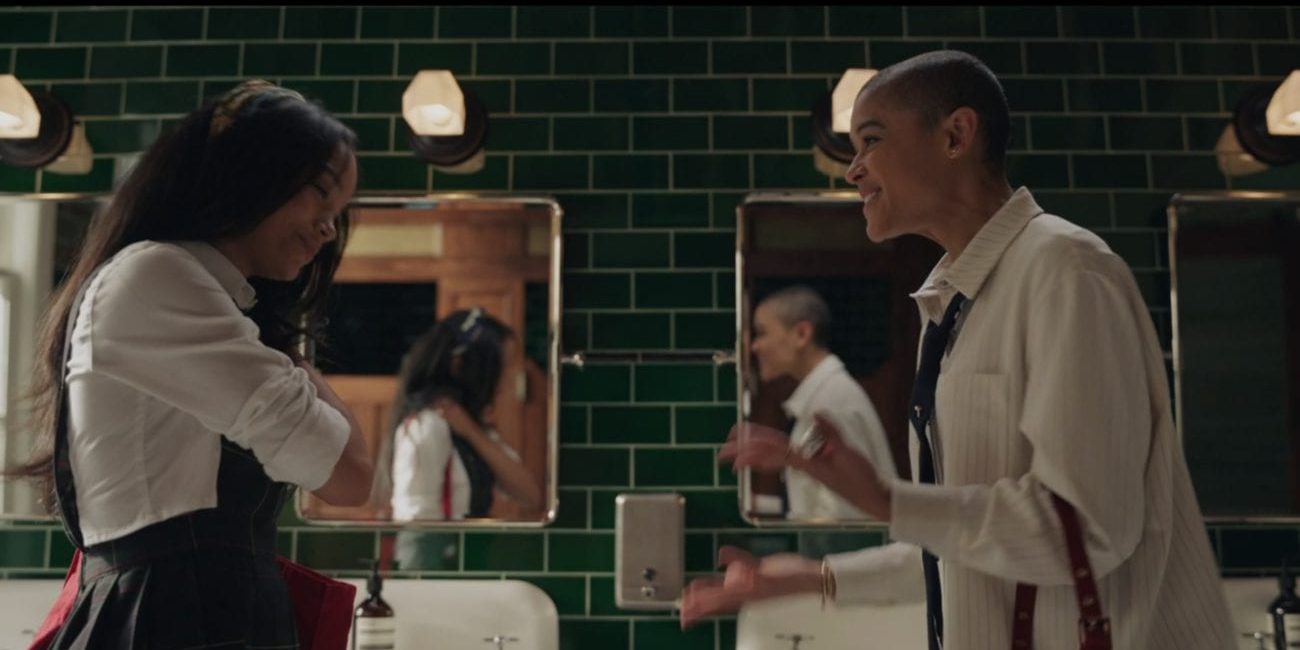 Zoya, left, a black girl with long, straightened dark hair wearing a headband, white dress shirt, and a black overall skirt in the dark green-tiled school bathroom with her sister, Julien, a lighter-skinned black girl with a buzzcut wearing an oversized white and striped dress shirt with a black tie. They are happy to see each other.