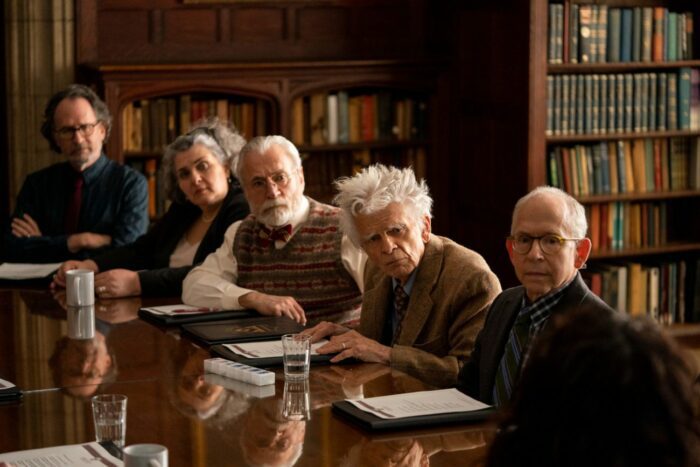 Several people, with Professor Renz (Bob Balaban) on the right sit at a table looking toward the camera, with bookshelves behind them