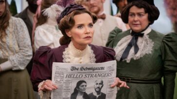 Mrs. Layton (Kristin Chenoweth) standing in the middle of a group of citizens holding a newspaper with a picture of Josh and Mel and the headline "Hello Strangers" on the front page and looking intently toward the crowd