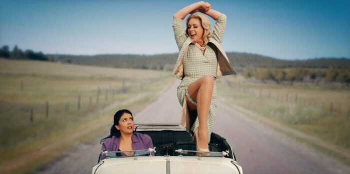 The Countess (Jane Krakowski) stands on the top of her steering wheel dancing as Mel (Cecily Strong) looks on