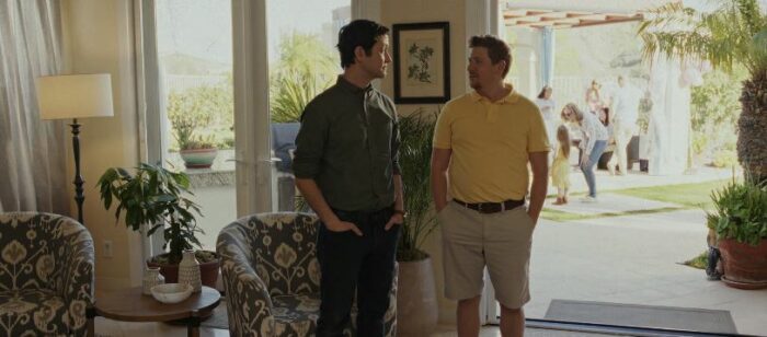 Josh and Aaron stand with their hands in their pockets at the party, in a room that opens to the outdoors behind them