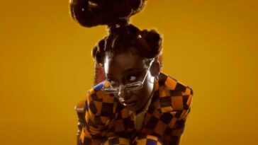 Sometimes I Might Be Introvert: Little Simz sits hunched over shyly in a small chair, wearing a noisy chequered suit that matches the backdrop. Her hair is magnificent.