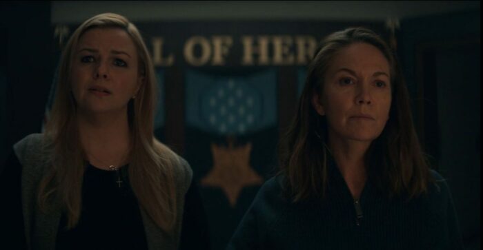 Kimberley Cunningham (Amber Tamblyn) and President Blake (Diane Lane) stand in a darkened room in front of a sign that says "Hall of Heroes" staring straight ahead